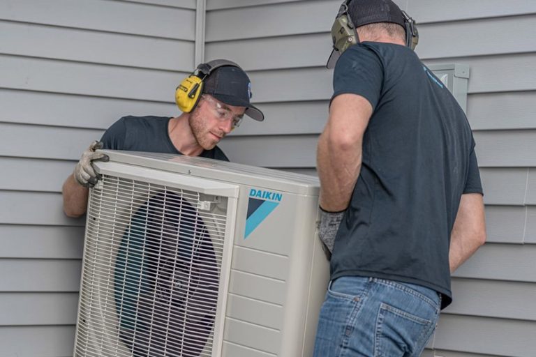 Two HVAC technicians installing an outdoor Daikin HVAC unit next to a light gray home with vinyl siding.